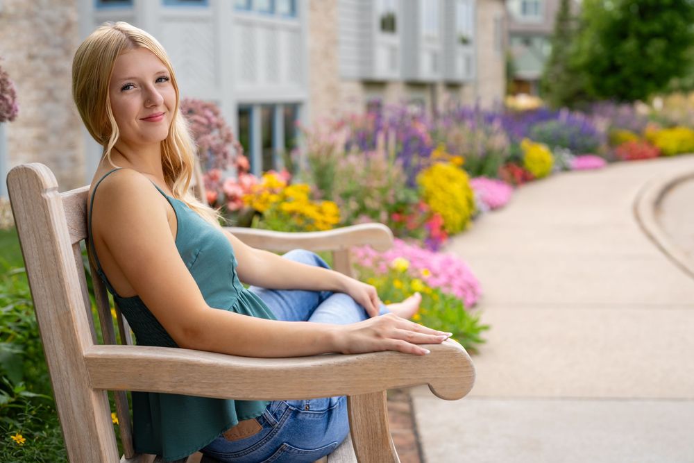 Young blonde woman sitting on a wooden bench surrounded by colorful garden flowers outdoors.
