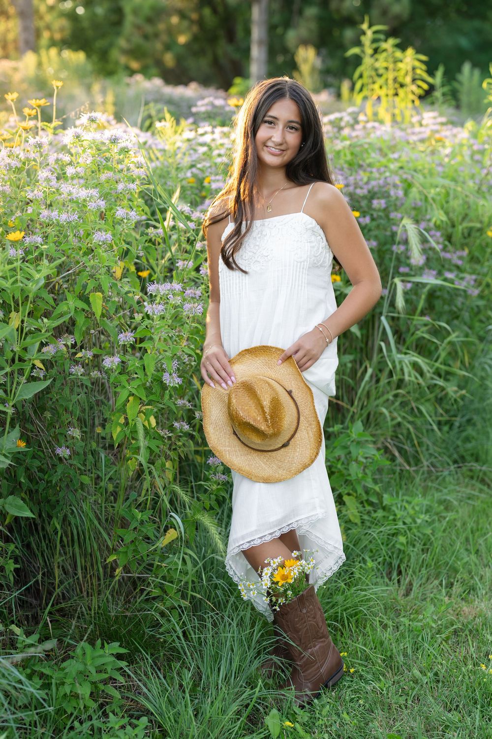 Young woman in white sundress holding straw hat and wildflowers, standing in lush green meadow wearing brown cowboy boots.