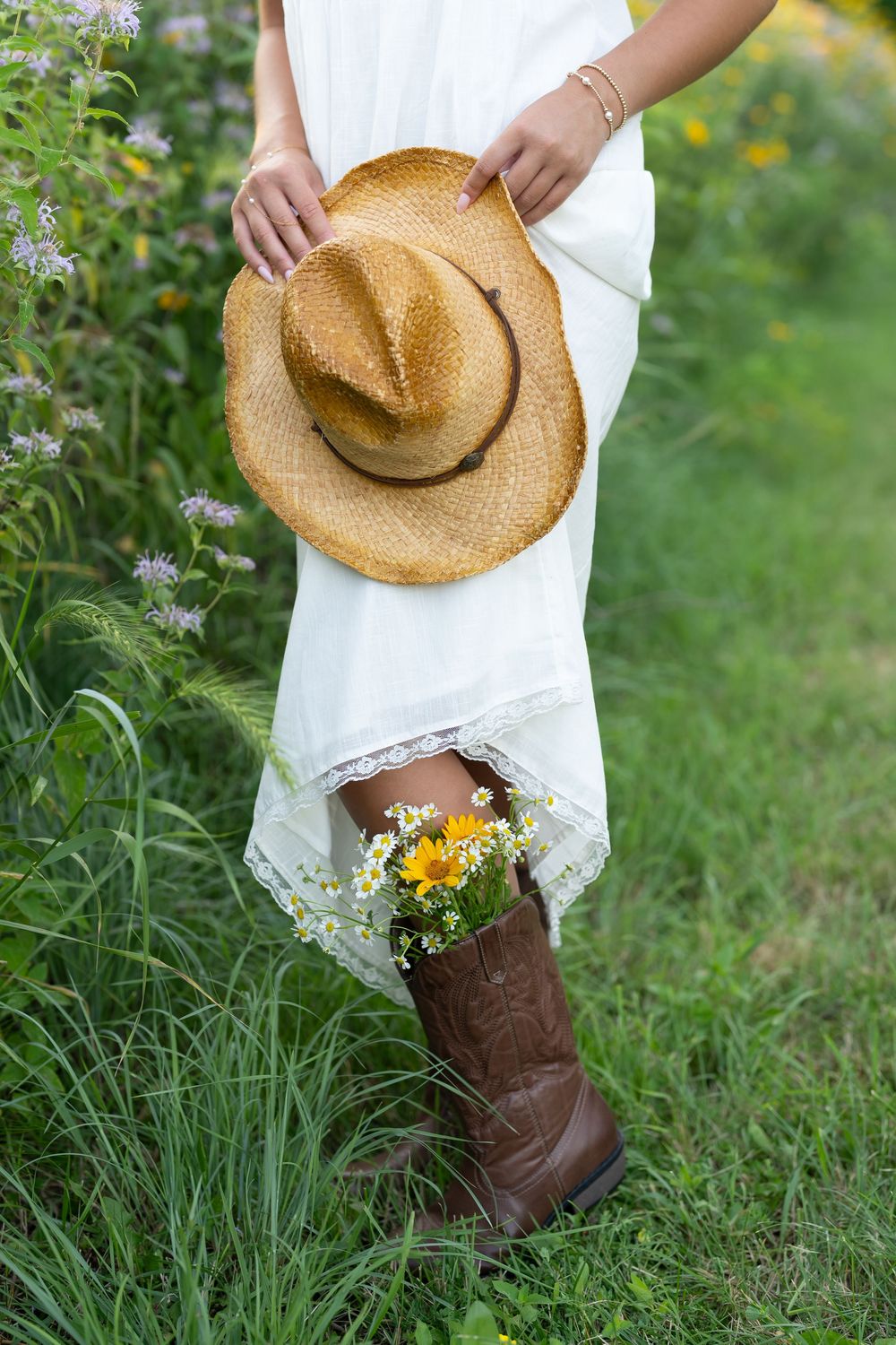 Woman in white dress holding straw hat with wildflowers tucked into brown cowboy boot in green field.