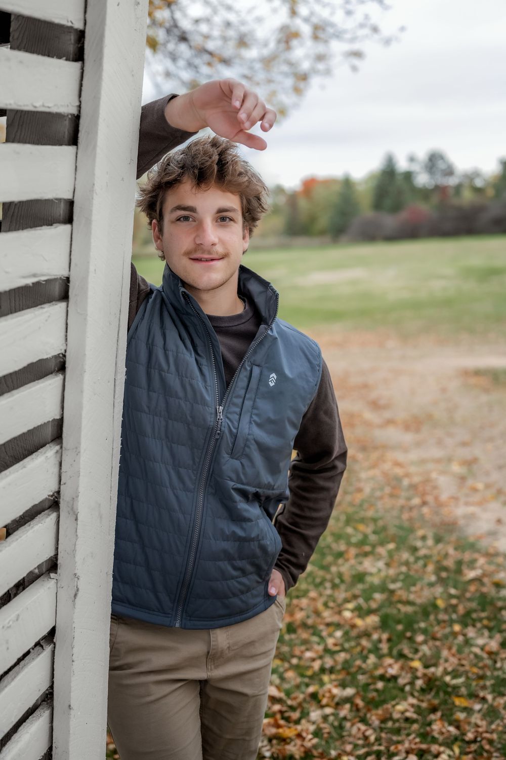 Young man in a blue quilted vest leaning against a white barn wall surrounded by autumn foliage.