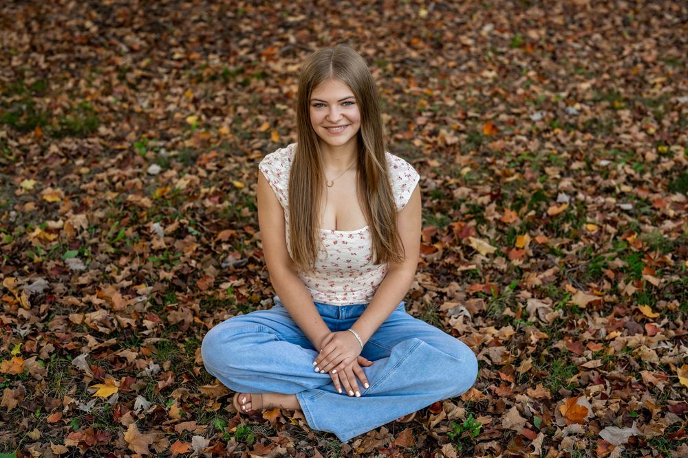 Young woman with long hair sits cross-legged on autumn leaves, smiling, wearing a floral top and blue jeans.