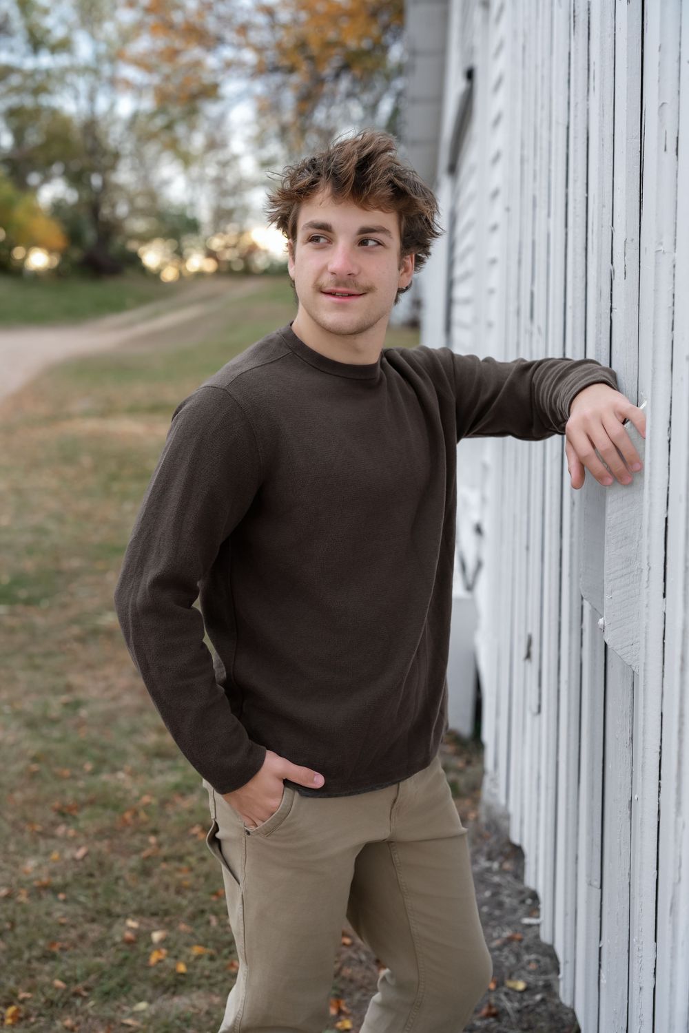 Young man in brown sweater and khaki pants leaning against white barn wall outdoors in autumn.
