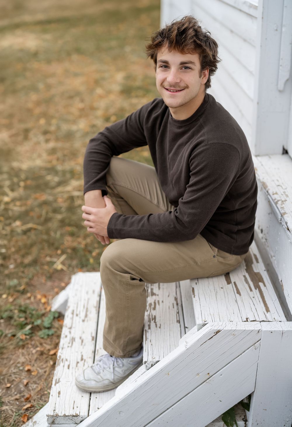 Young man in brown sweater and khaki pants smiling while sitting on white wooden steps outdoors in autumn.