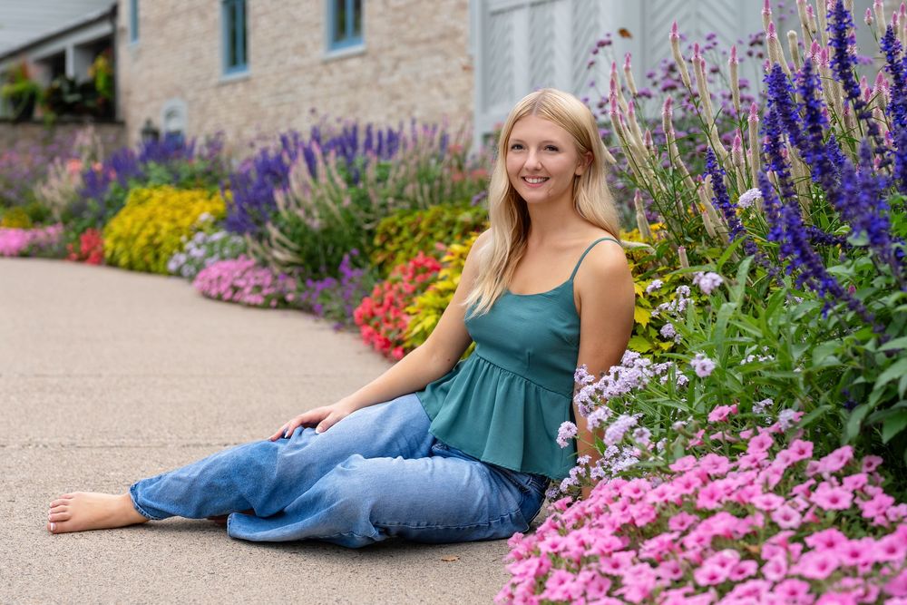 Young blonde woman sitting barefoot among vibrant colorful garden flowers in jeans and green top.