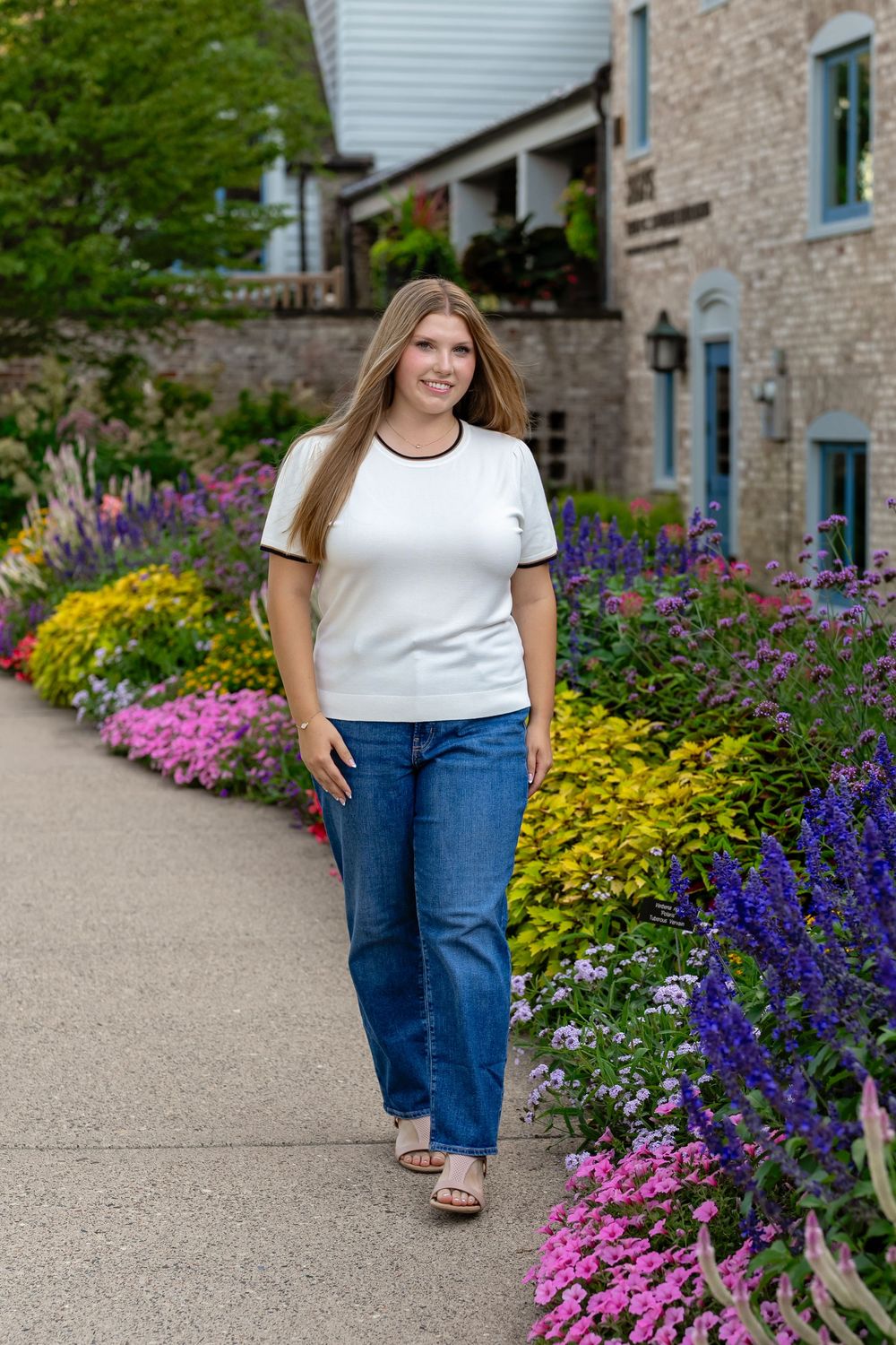 Woman in white top and jeans walking along a colorful garden path beside a stone building.