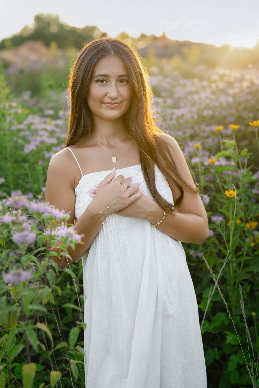 Young woman in white sundress standing in wildflower field at golden hour sunset, smiling softly at camera.