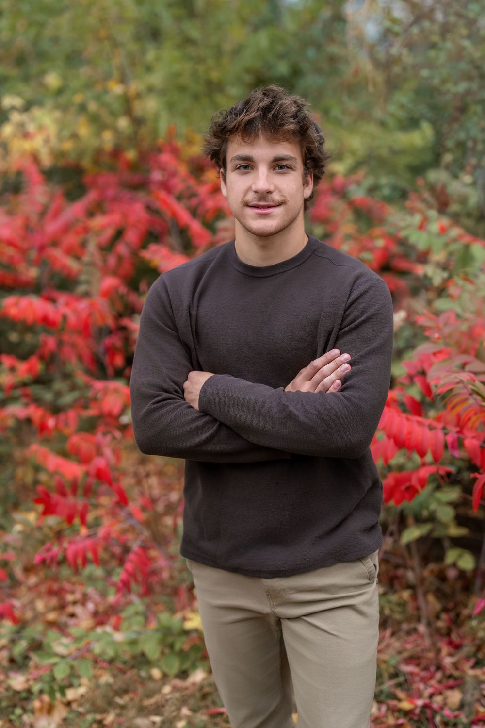 Young man with curly hair in brown sweater poses arms crossed before vibrant red autumn foliage.
