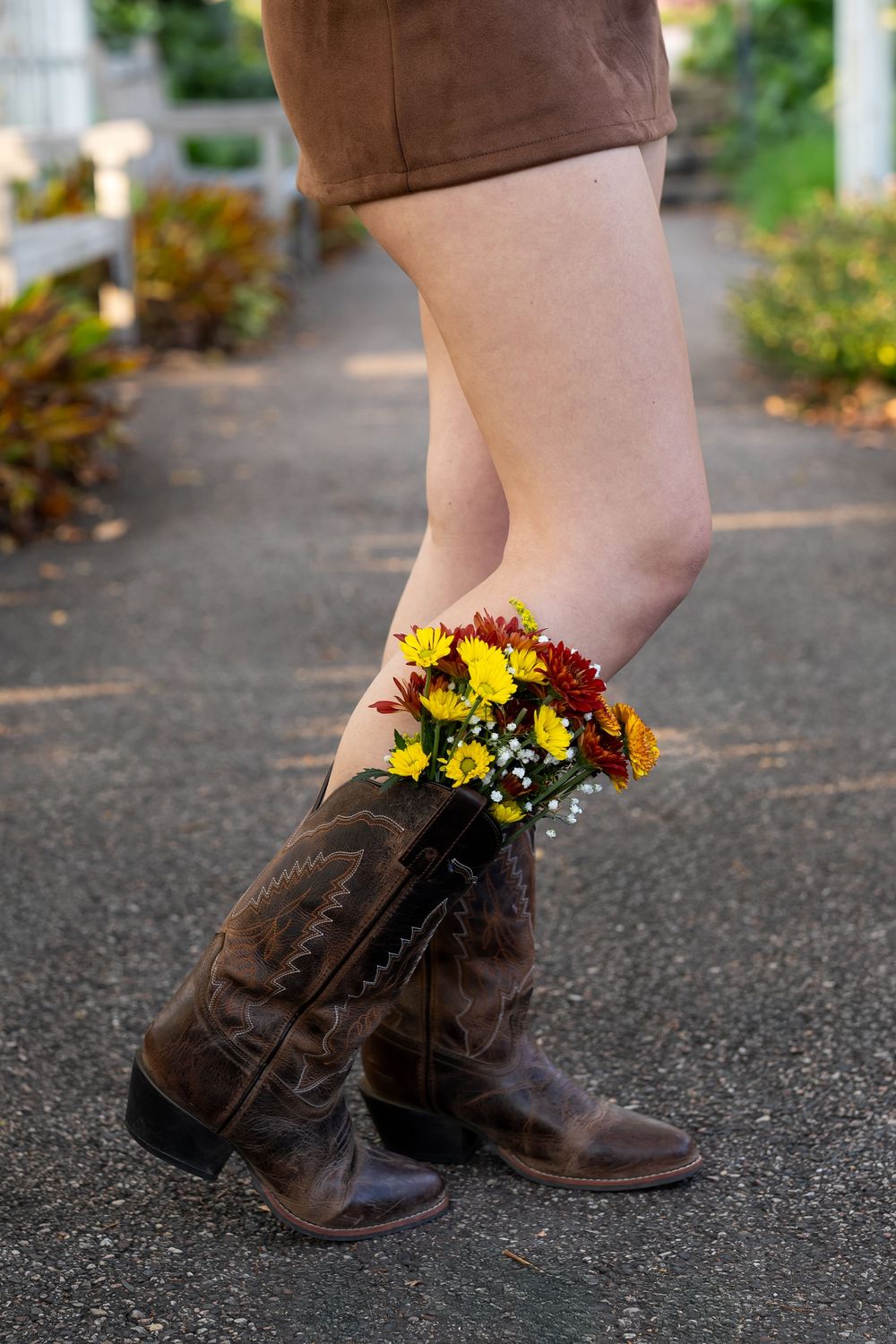 Woman wearing brown cowboy boots with colorful yellow and red wildflowers tucked inside, standing on a path outdoors.