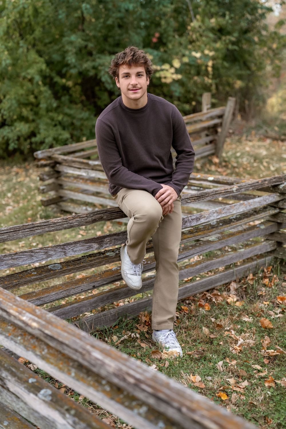 Young man in brown sweater and khaki pants sitting on a rustic wooden fence outdoors in autumn.