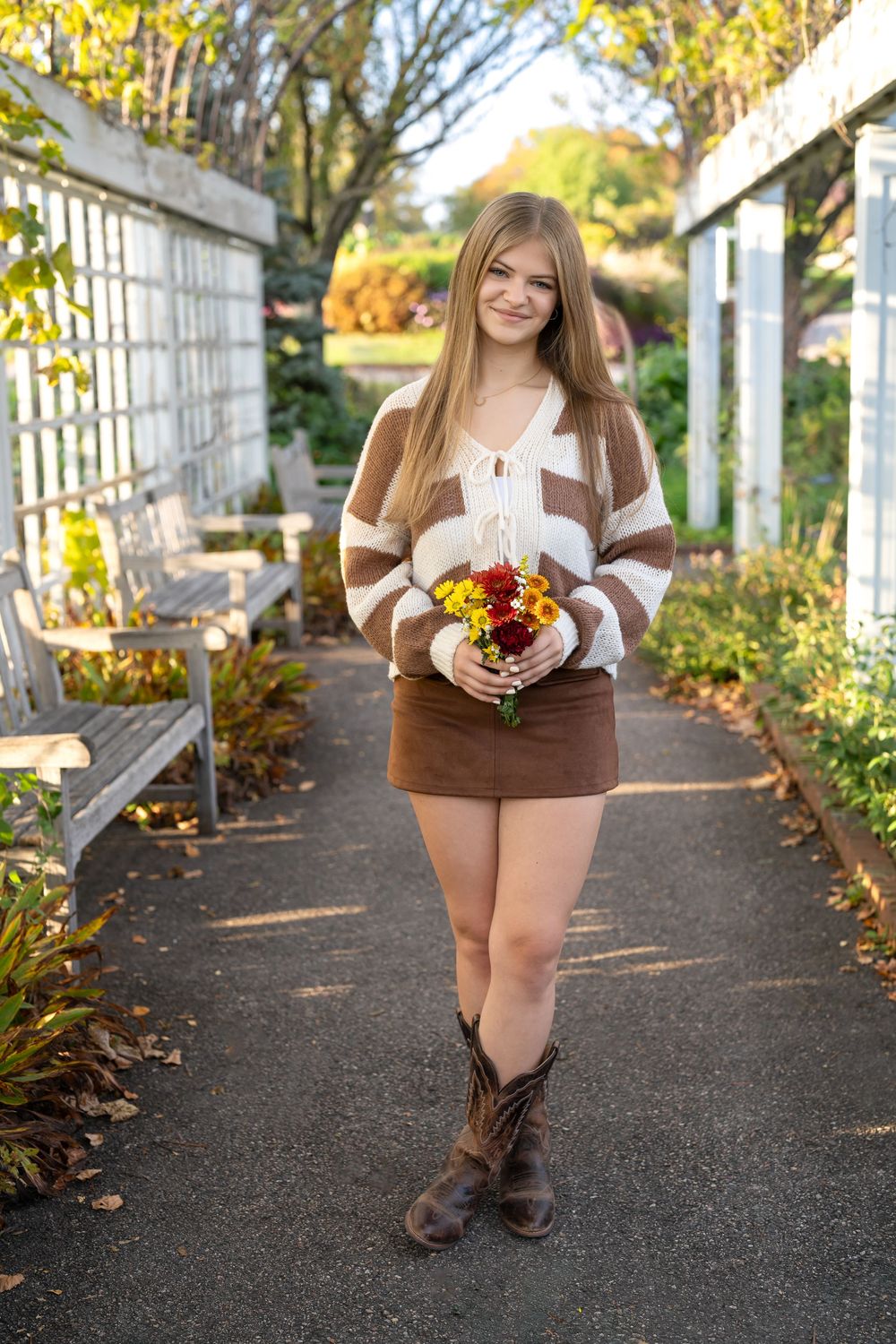 Young woman in striped sweater, brown mini skirt, and cowboy boots holding colorful flowers in an autumn garden path.