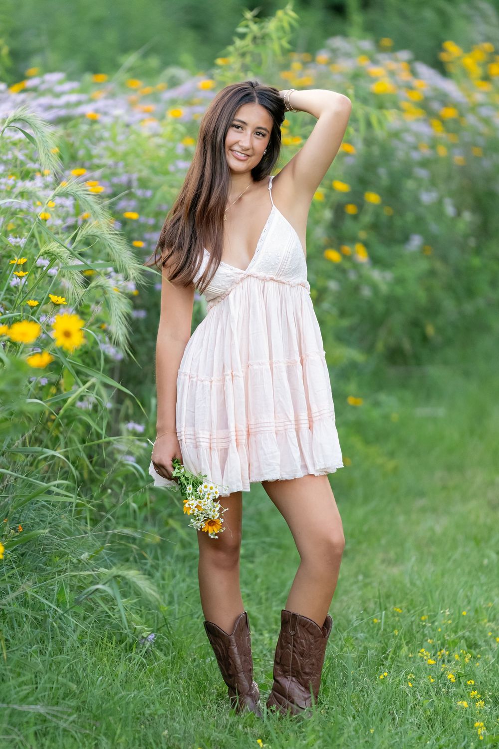 Young woman in white sundress and cowboy boots holding wildflowers, smiling in a garden with yellow blooms.