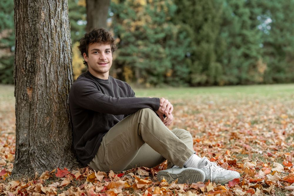 Young man smiling, seated against a tree trunk surrounded by autumn leaves in a park setting.