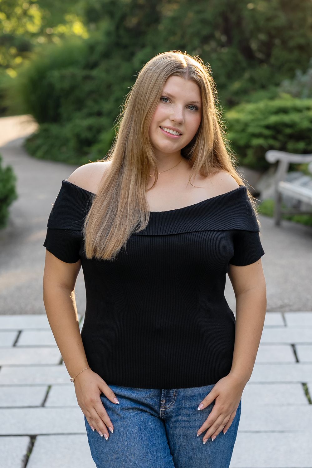 A smiling blonde woman in a black off-shoulder top and jeans poses on a stone path in a lush green park.