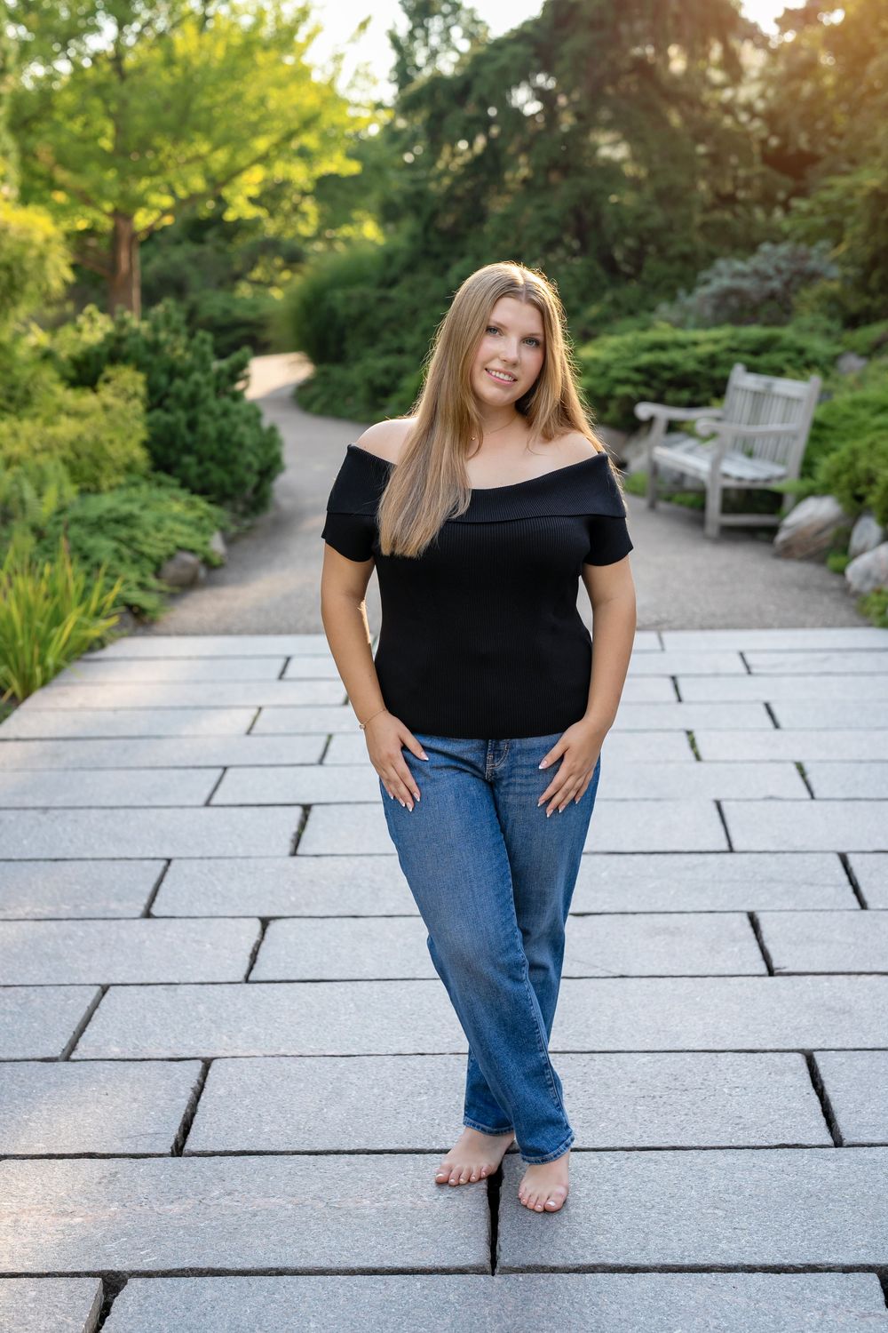 Woman in black off-shoulder top and blue jeans standing barefoot on stone path in lush green garden.
