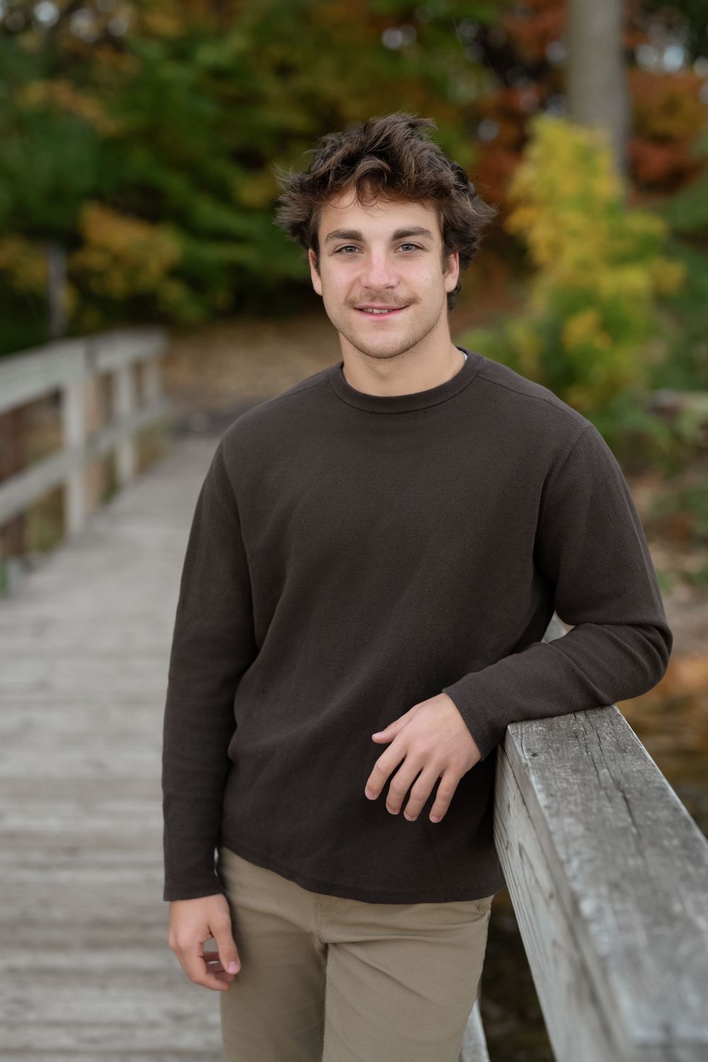 Young man in brown long-sleeve shirt leaning on wooden bridge railing with autumn foliage background.