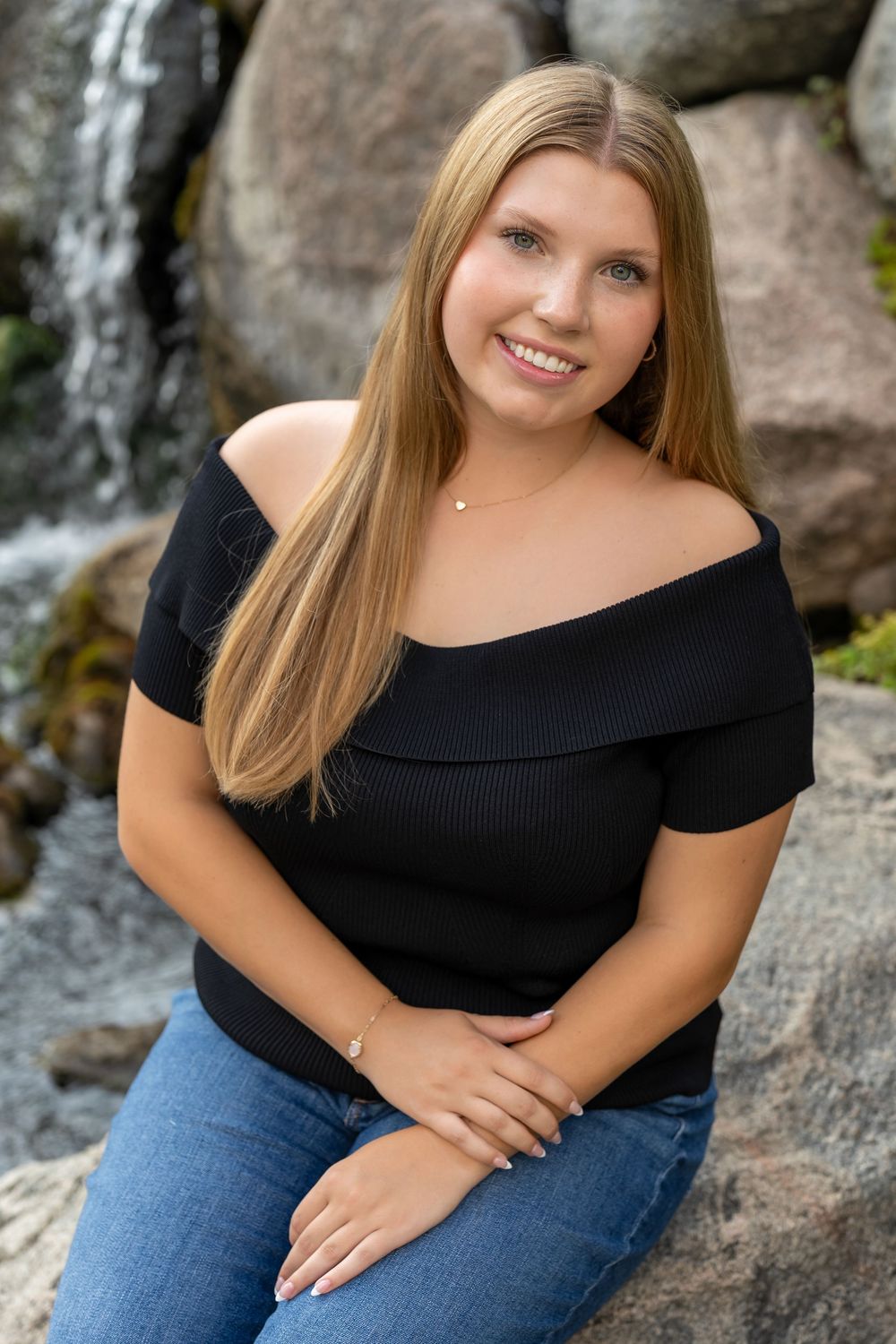 Young woman with long blonde hair smiling, wearing a black off-shoulder top and jeans, seated on rocks near a waterfall.