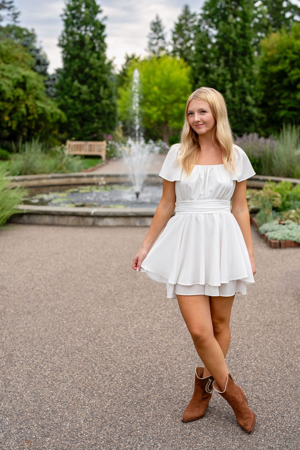 Young blonde woman in white mini dress and brown cowboy boots posing in a garden with fountain.