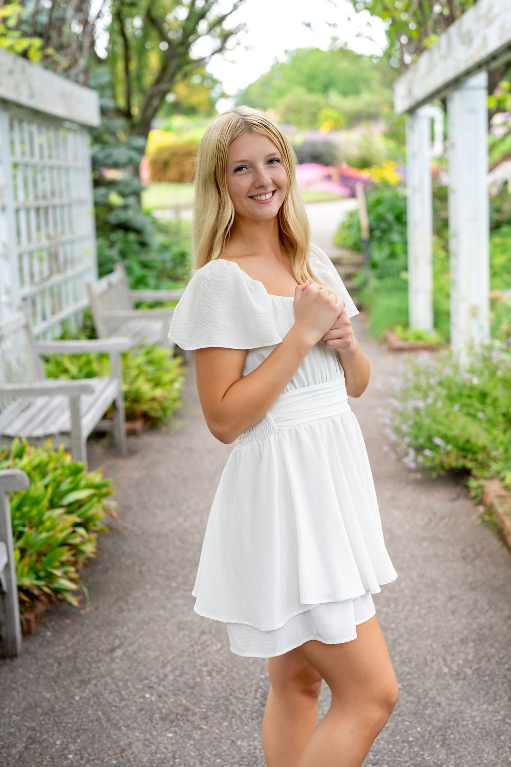 Smiling blonde woman in white flutter-sleeve mini dress posing in a lush garden pathway with wooden trellis.