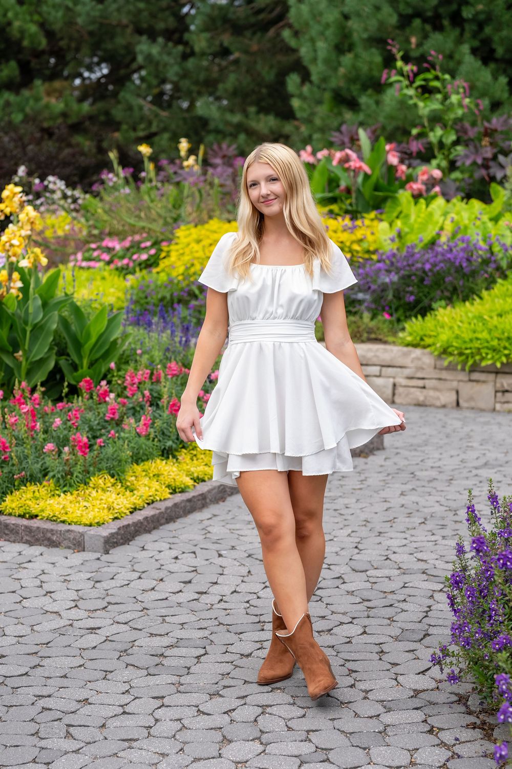 Young blonde woman in white mini dress and brown boots smiling in a colorful garden.