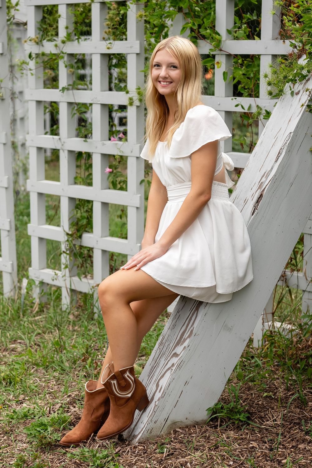 Smiling blonde woman in white dress and brown cowboy boots sitting against white garden trellis outdoors.