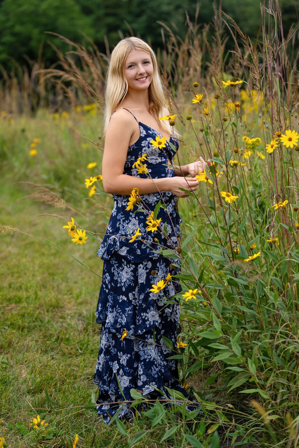 Smiling blonde woman in a navy floral maxi dress standing among yellow wildflowers in a lush green field.