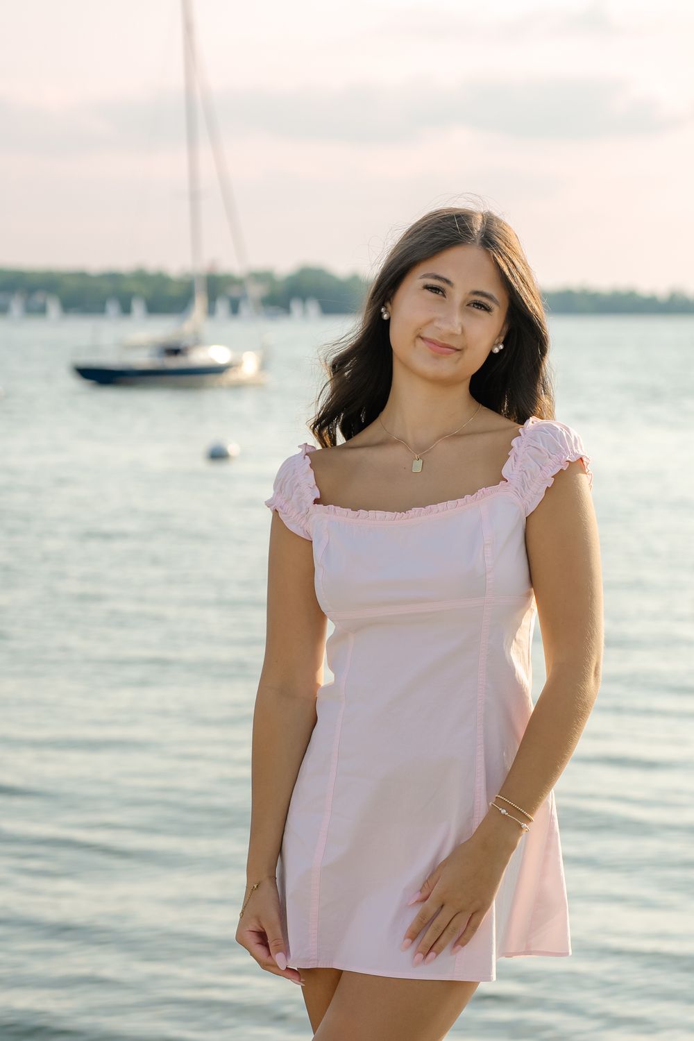 Young woman in pink mini dress poses by waterfront with sailboat in background on a calm evening.