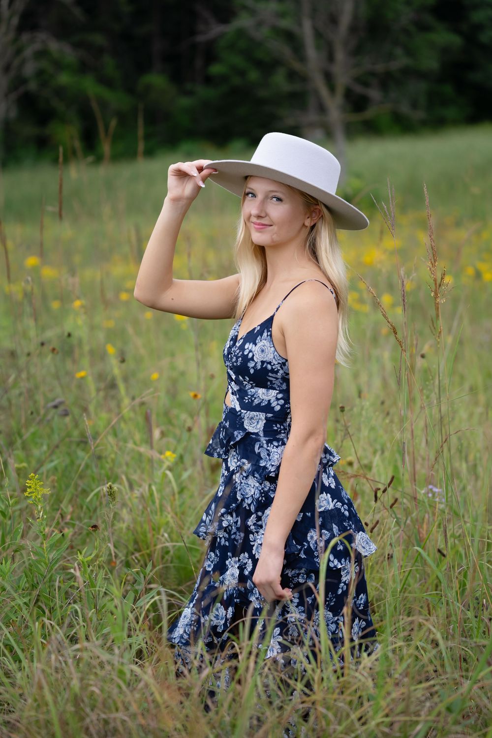 Young blonde woman in floral dress and white hat posing gracefully in a wildflower meadow.