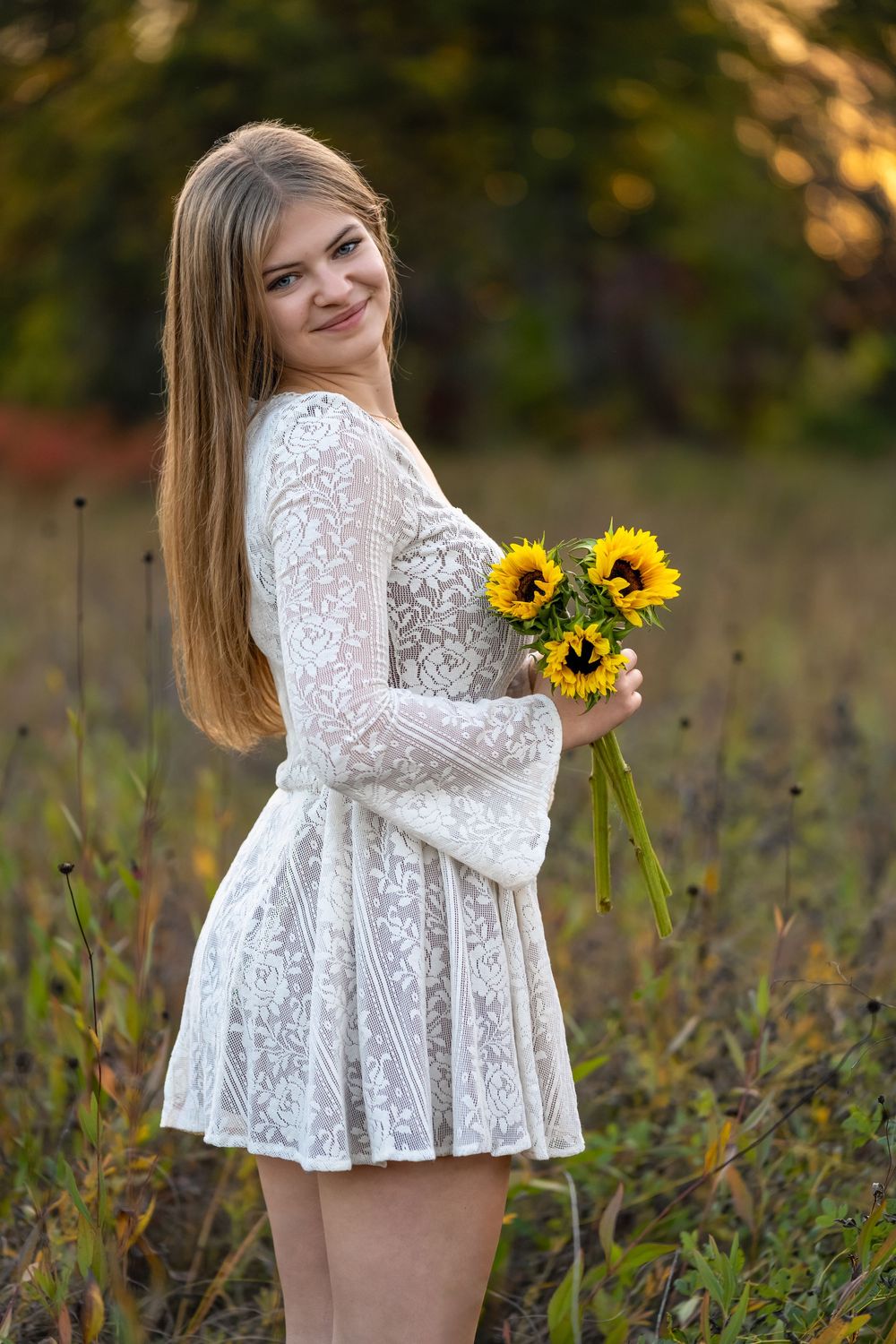 Young woman in white lace dress holding sunflowers smiling outdoors in a field during golden hour.