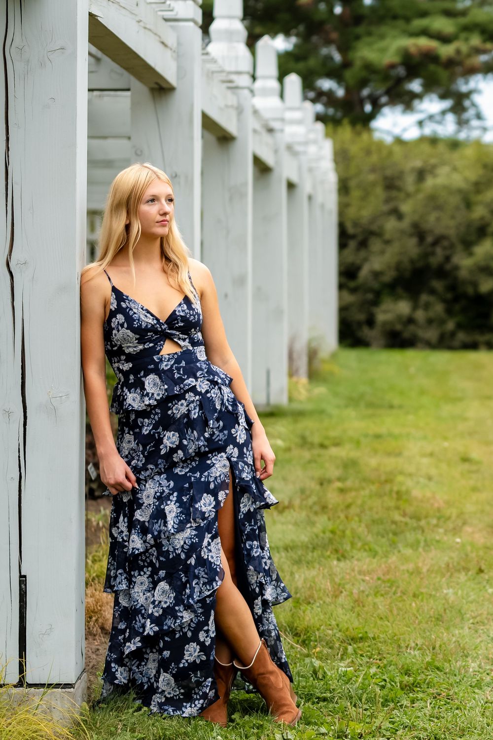 Blonde woman in navy floral maxi dress and brown boots leaning against white wooden columns outdoors.