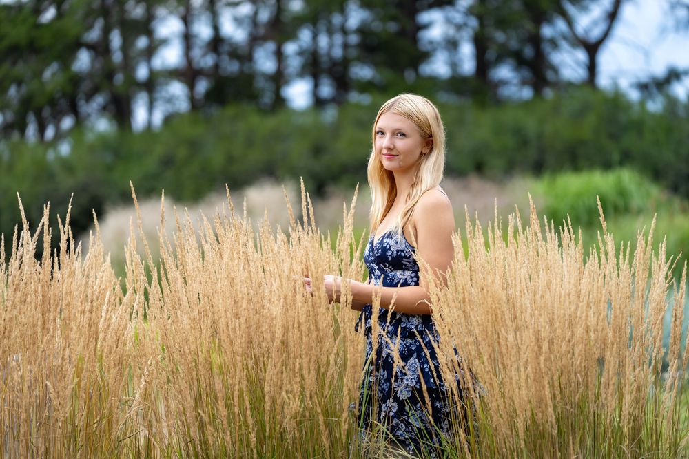 Young blonde woman in floral dress standing in golden tall grass field with green trees in background.