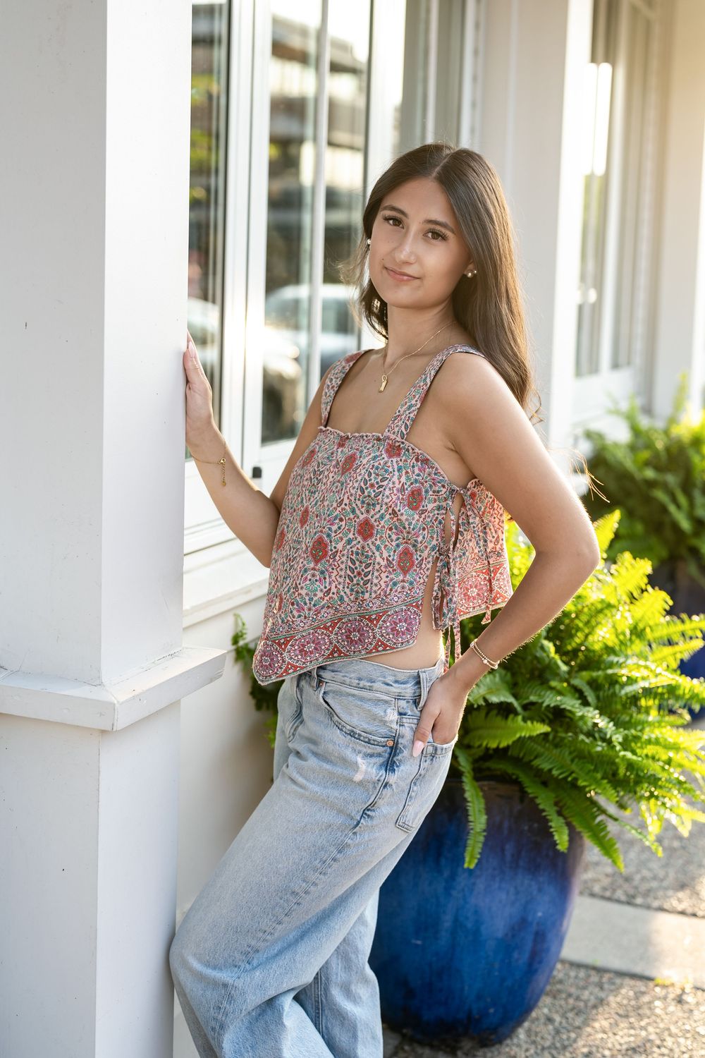 Young woman in floral crop top and jeans posing by white column with green ferns outdoors.
