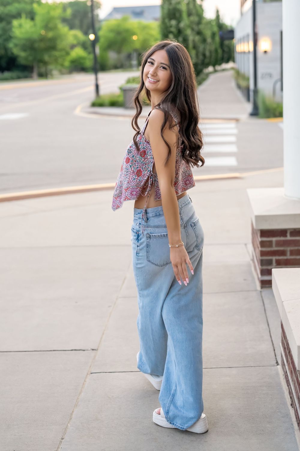 Young woman in floral crop top and wide-leg jeans smiling over shoulder on urban sidewalk.