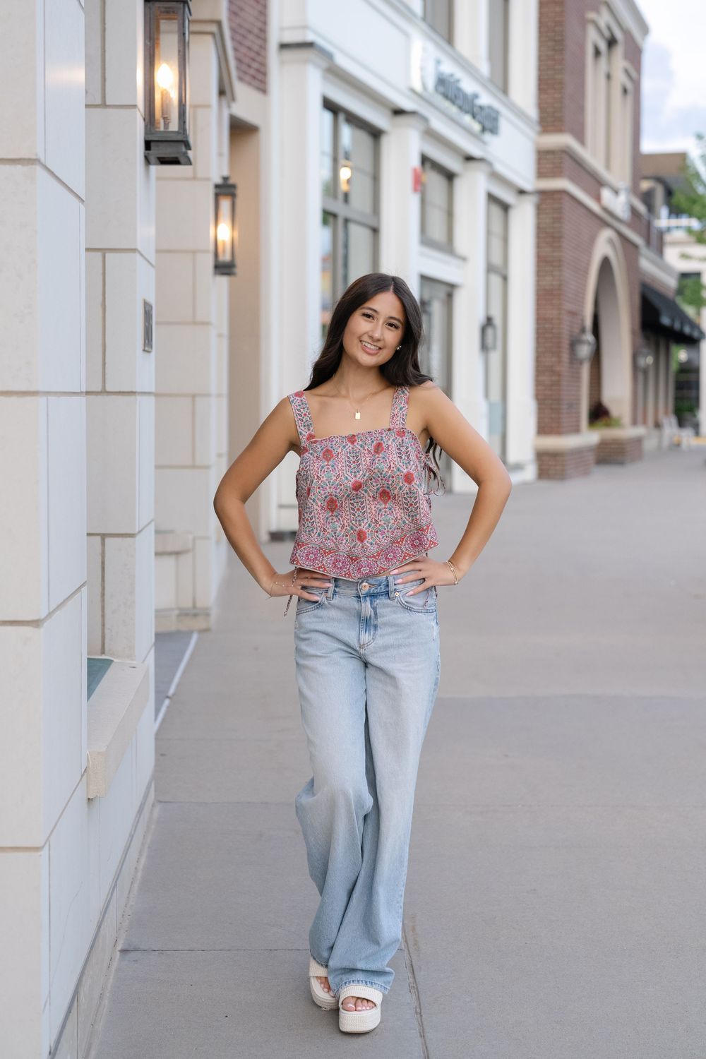 Young woman in floral crop top and wide-leg jeans poses on a sidewalk outside retail shops.
