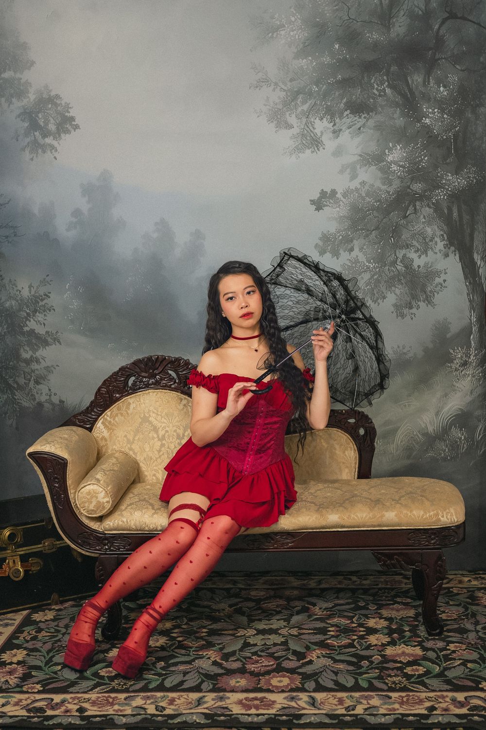 Young woman in red velvet dress and stockings holding black lace parasol on vintage chaise lounge.