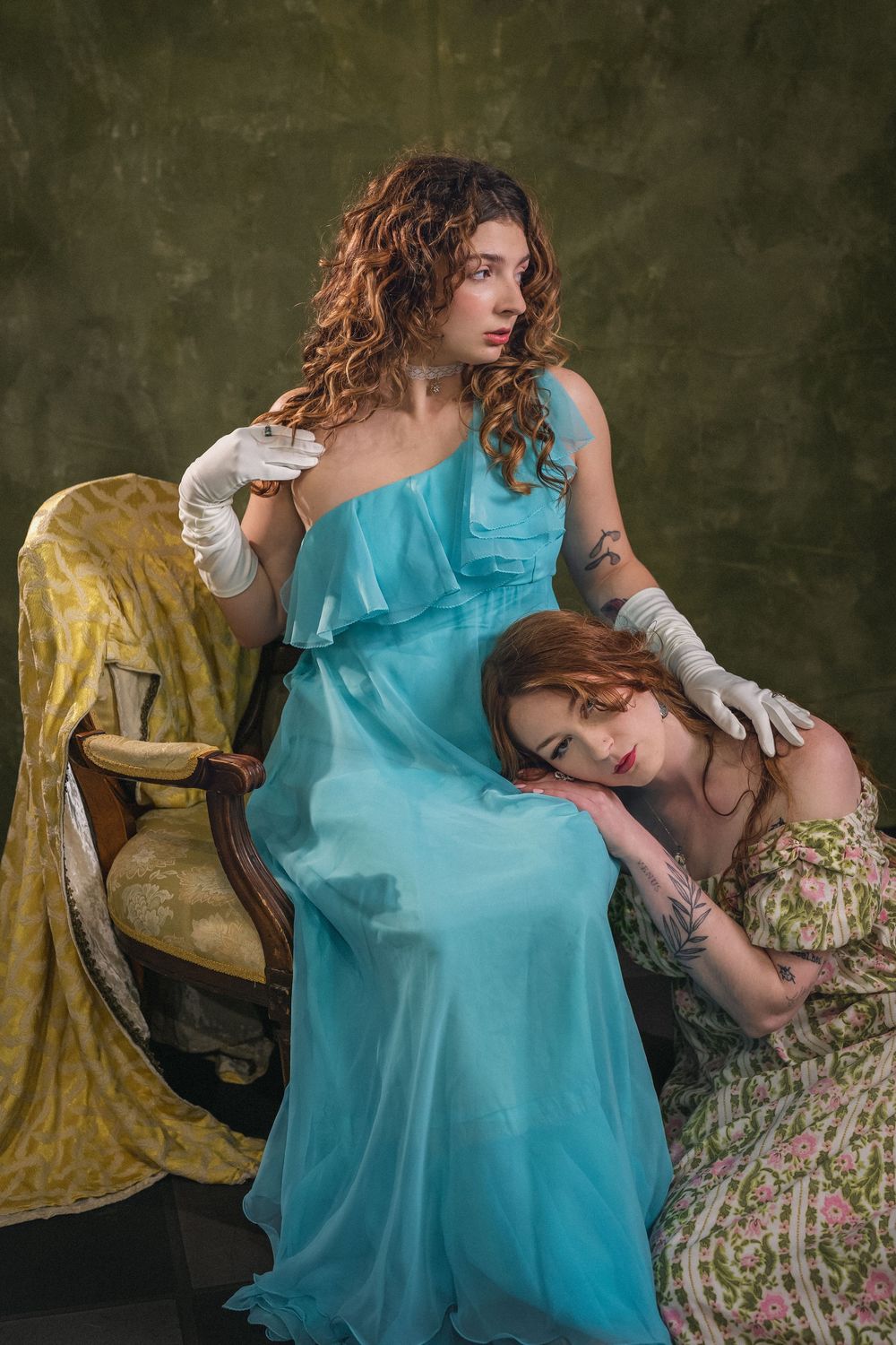Two women in vintage gowns posing together, one in blue dress with white gloves, other in floral dress.