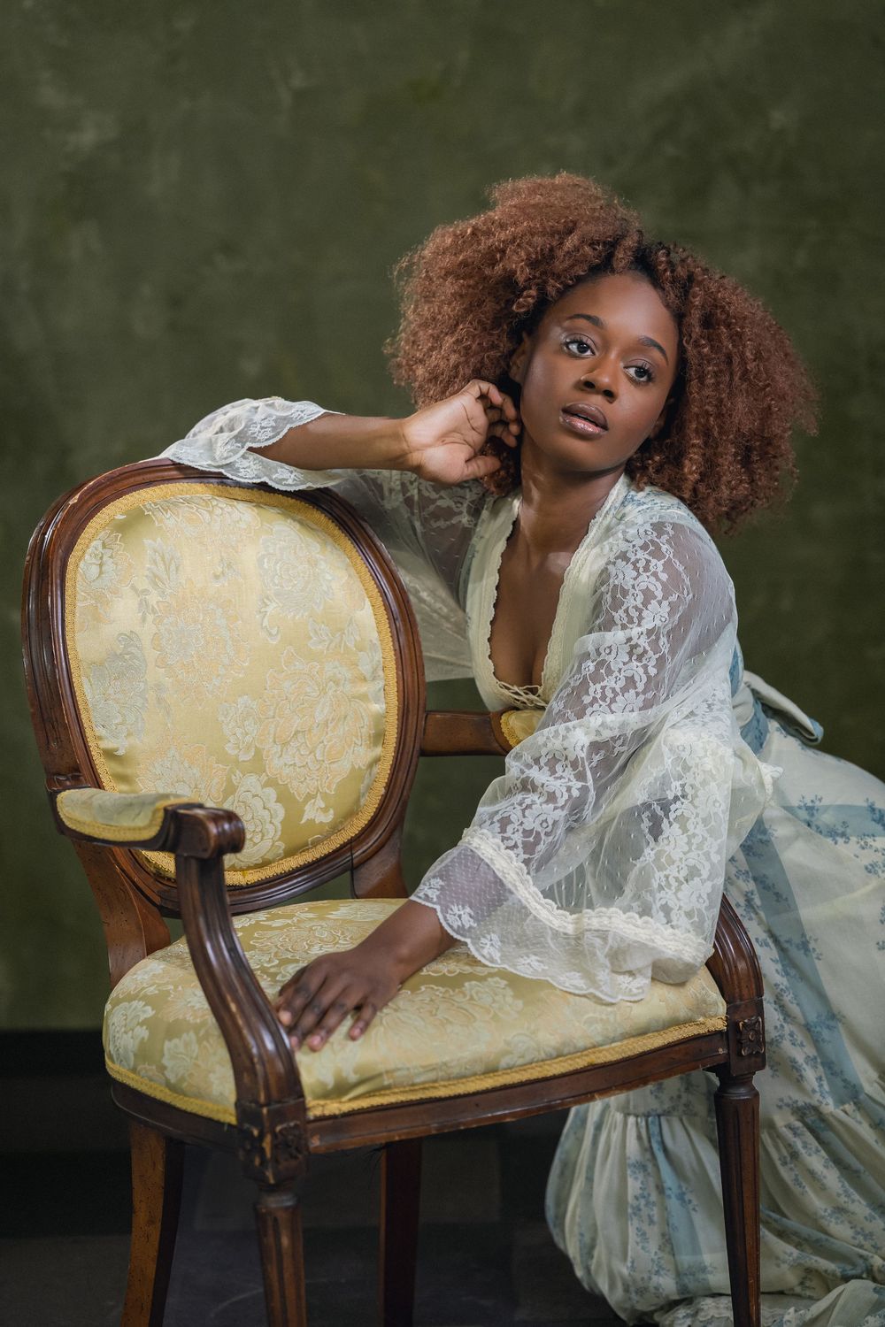 Black woman with curly auburn hair in white lace robe leaning on ornate gold vintage chair.