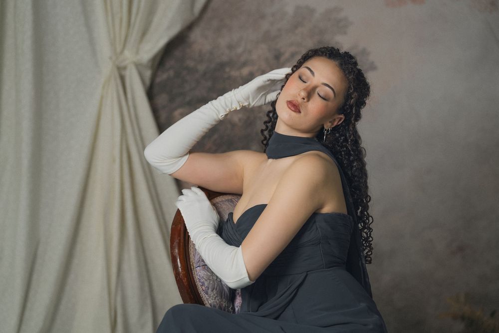Graceful woman in gray strapless dress and white gloves posing dramatically with eyes closed, studio setting.