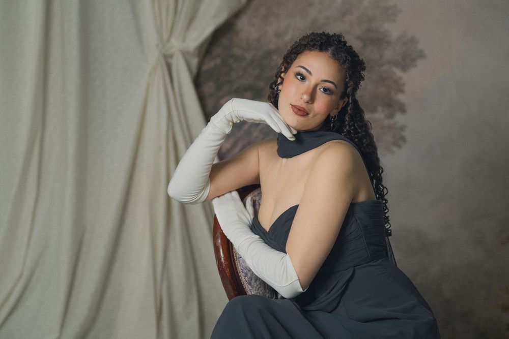 Elegant woman in gray strapless gown with white gloves seated on chair against vintage backdrop.