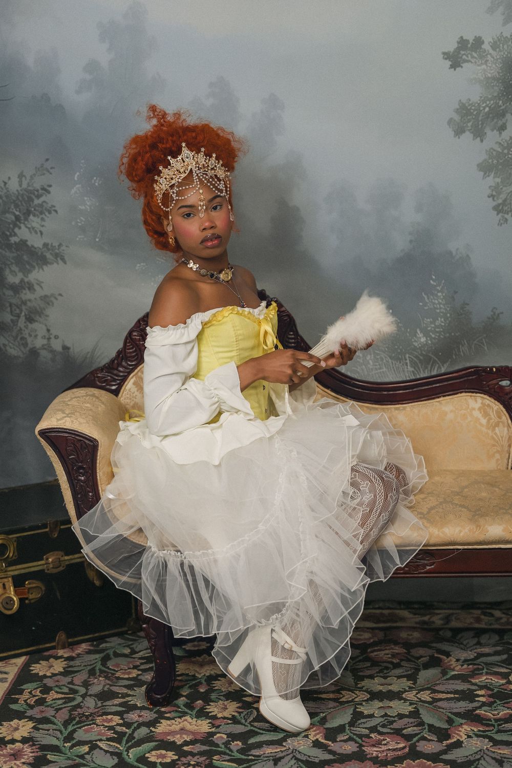 Young Black woman in yellow corset gown holding white feather fan seated on ornate vintage sofa.