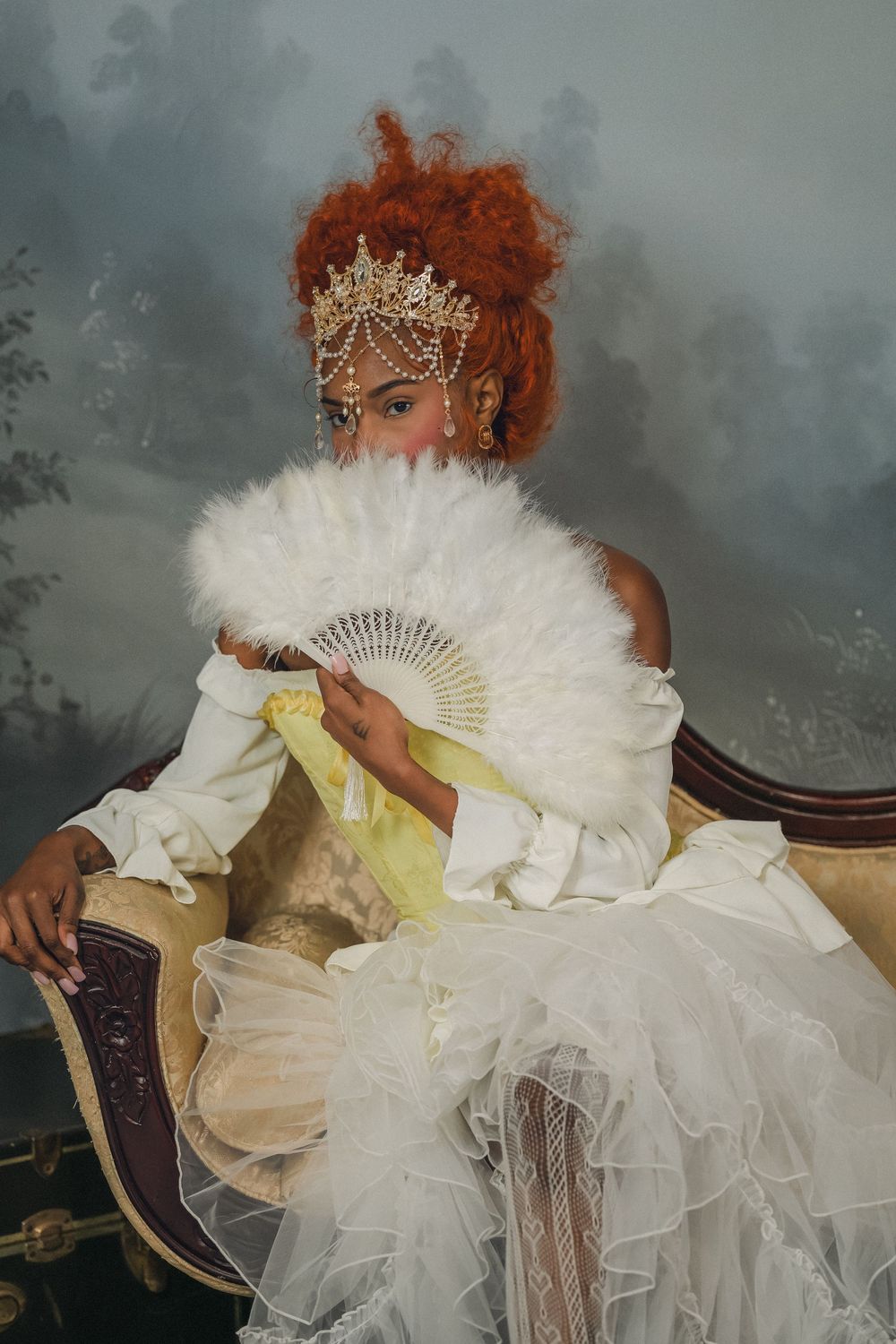 Black woman in white tulle gown with gold crown holding feather fan on antique sofa.