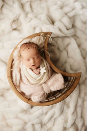 A sleeping newborn wrapped in soft white fabric lies peacefully in a wooden bowl against a white textured background.