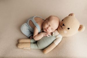 Newborn sleeping peacefully next to soft plush teddy bear on cream backdrop.