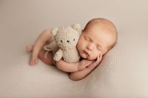 Sleeping newborn baby posed with soft teddy bear on neutral backdrop.