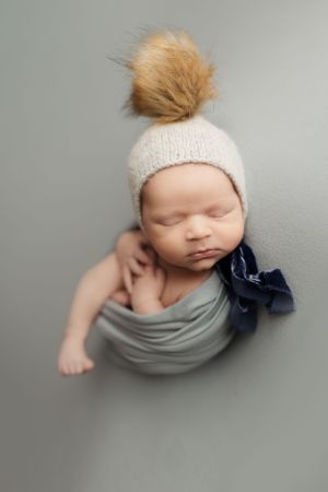 Newborn wearing knit headband with fur pom wrapped in gray fabric.
