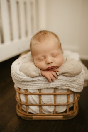 Sleeping newborn resting in white-lined wicker basket on dark wooden surface.