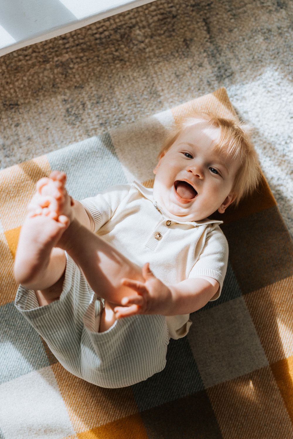 Happy laughing baby lying on plaid blanket holding feet up in sunny room.