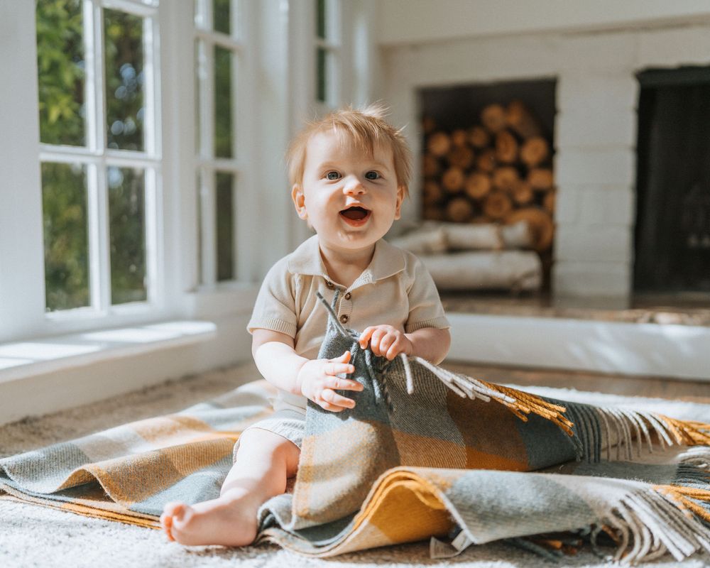 Smiling baby sitting on colorful plaid blanket near fireplace and large windows indoors.
