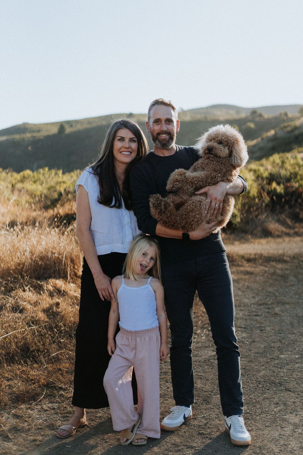 Happy family with young daughter and fluffy dog posing outdoors on a sunny hillside trail.