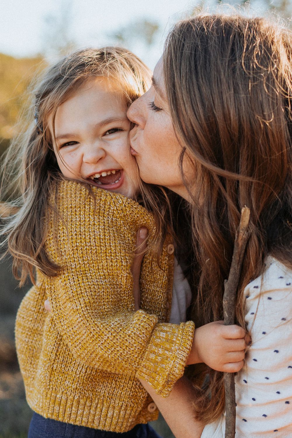 Mother kissing laughing young girl in yellow knit sweater outdoors in warm light.