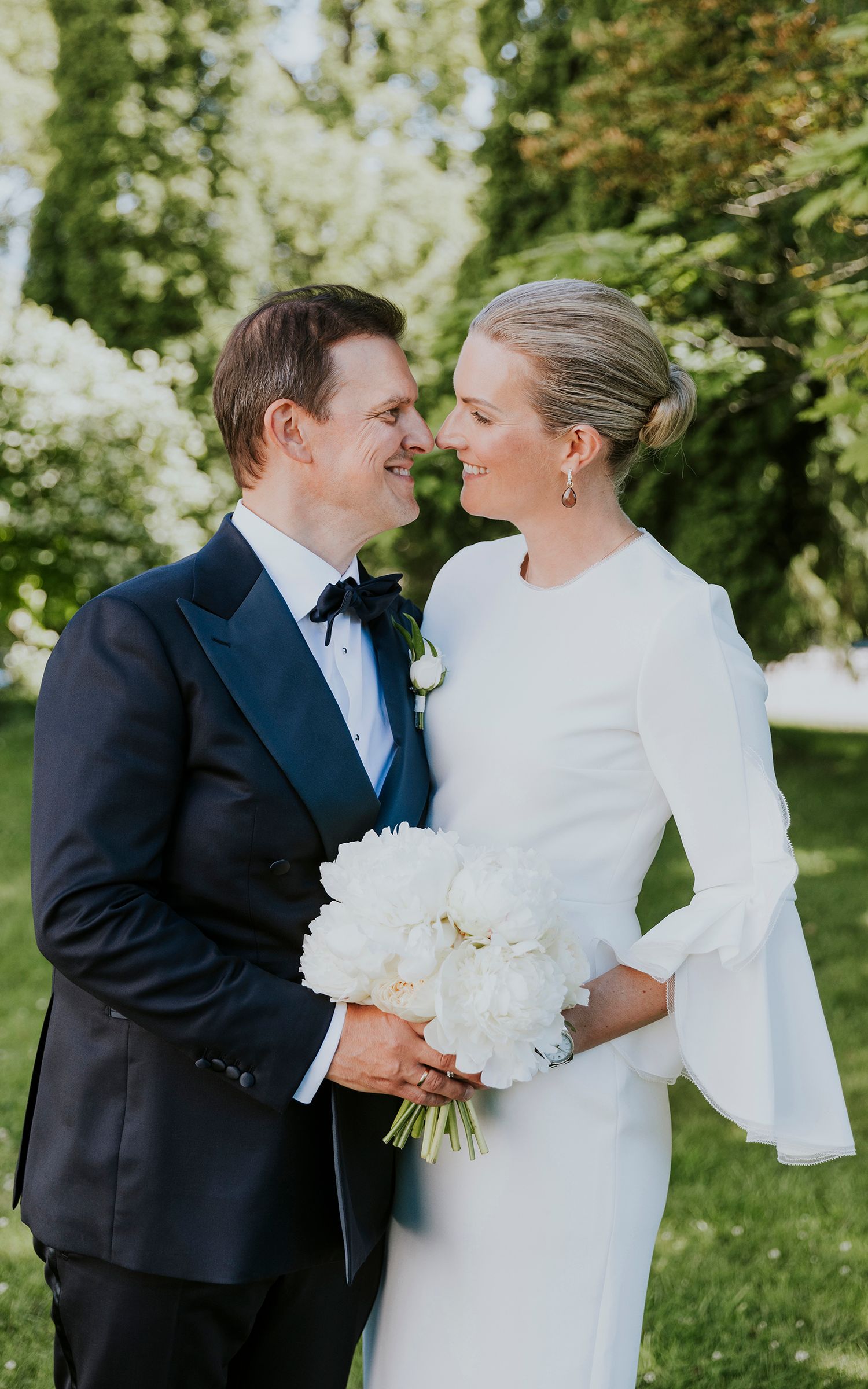 Brudepar som står rett og nesten kysser. Begge holder på blomsterbuketten som består av hvite peoner.